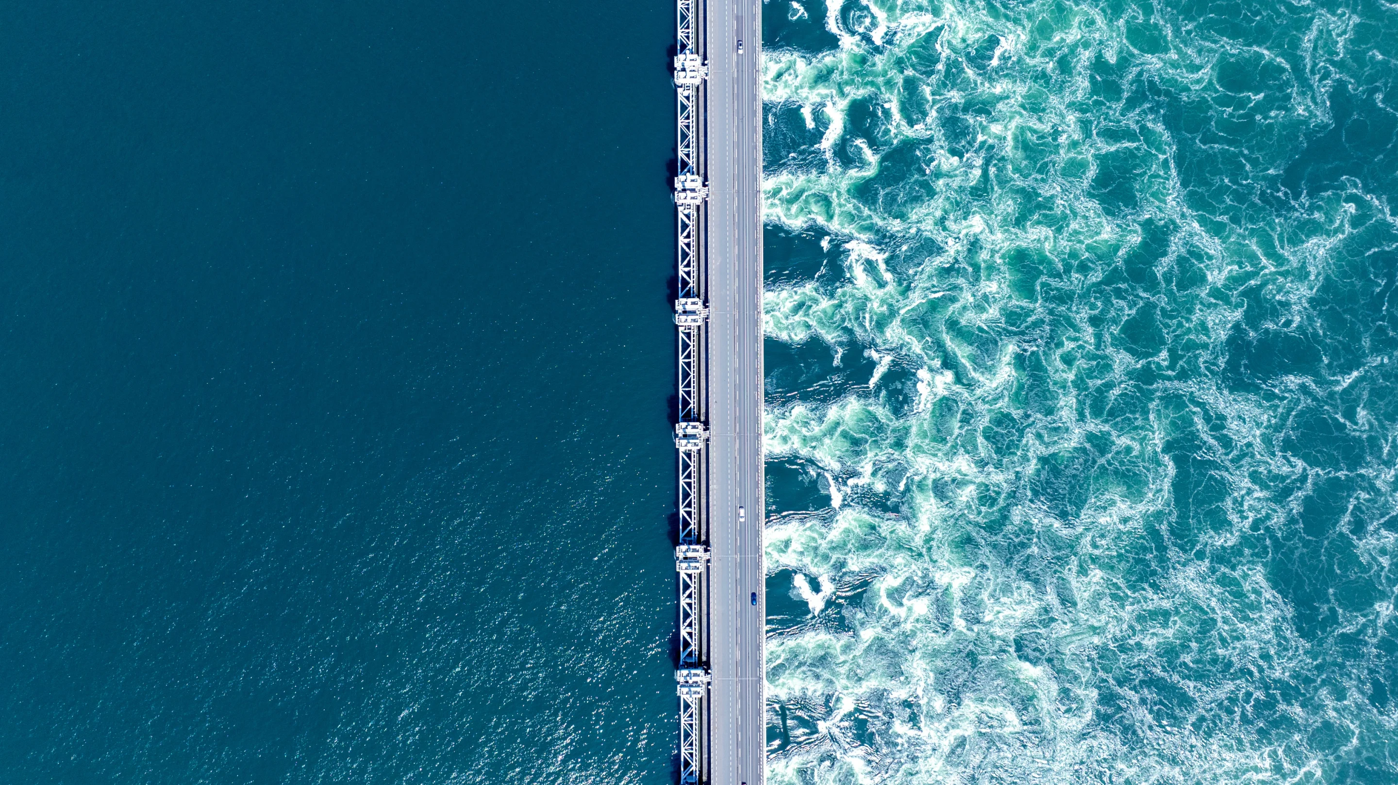 Aerial view of coastal infrastructure between calm water and turbulent waves, illustrating climate risk and infrastructure resilience.